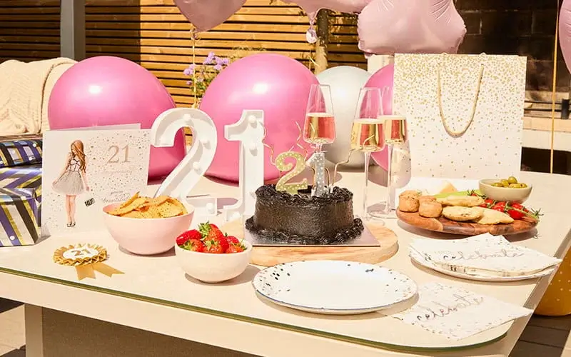 21st birthday celebration table with a chocolate cake, number candles, champagne glasses, pink balloons, snacks, and party decorations.
